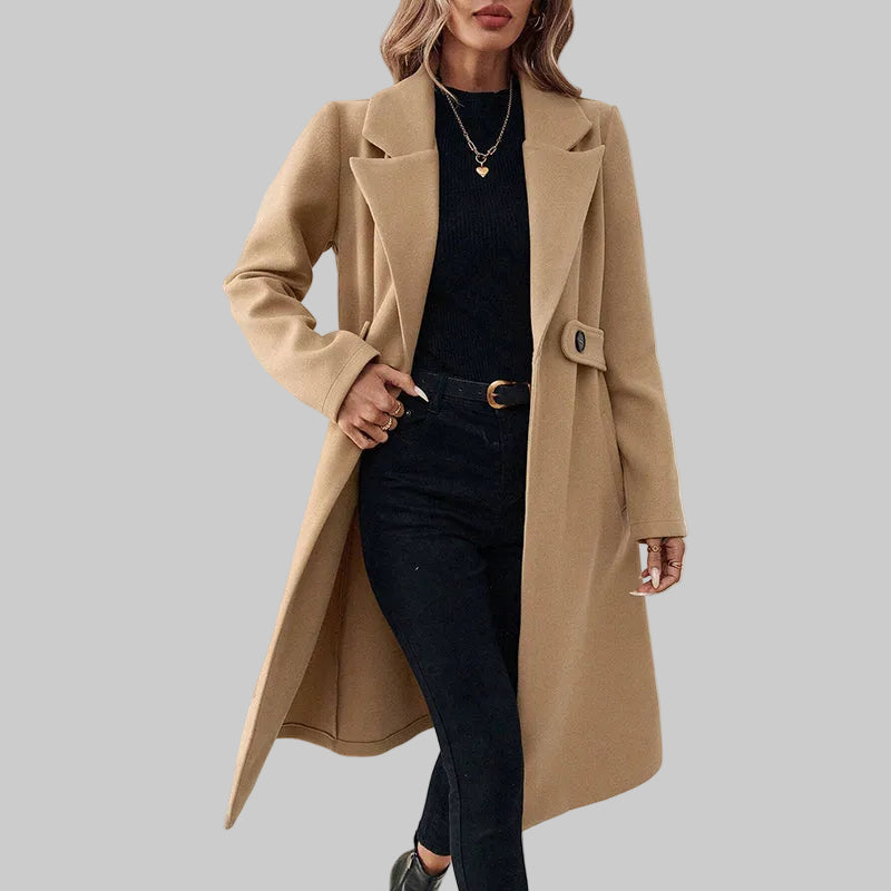 Woman wearing a beige coat over a black outfit in a minimalistic room.